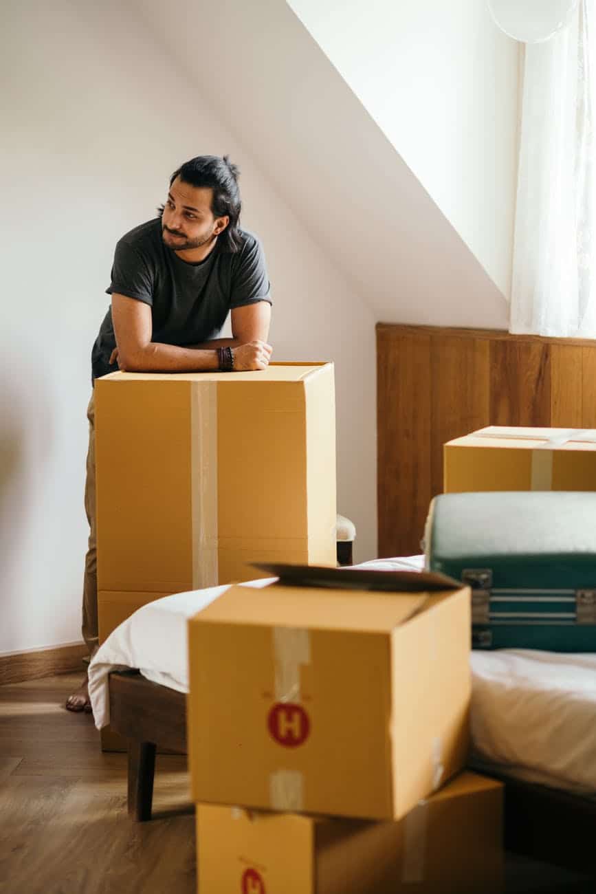 thoughtful ethnic man standing in new house leaning on unpacked carton box